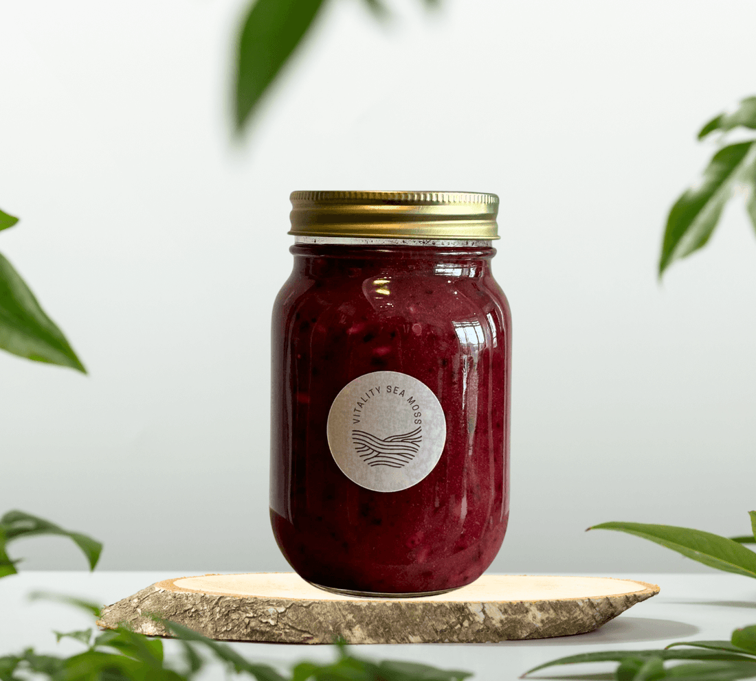 A glass jar of deep purple sea moss gel with a gold lid, labeled 'Vitality Sea Moss,' placed on a natural wood slab. Surrounded by green leaves and set against a light, minimal background.